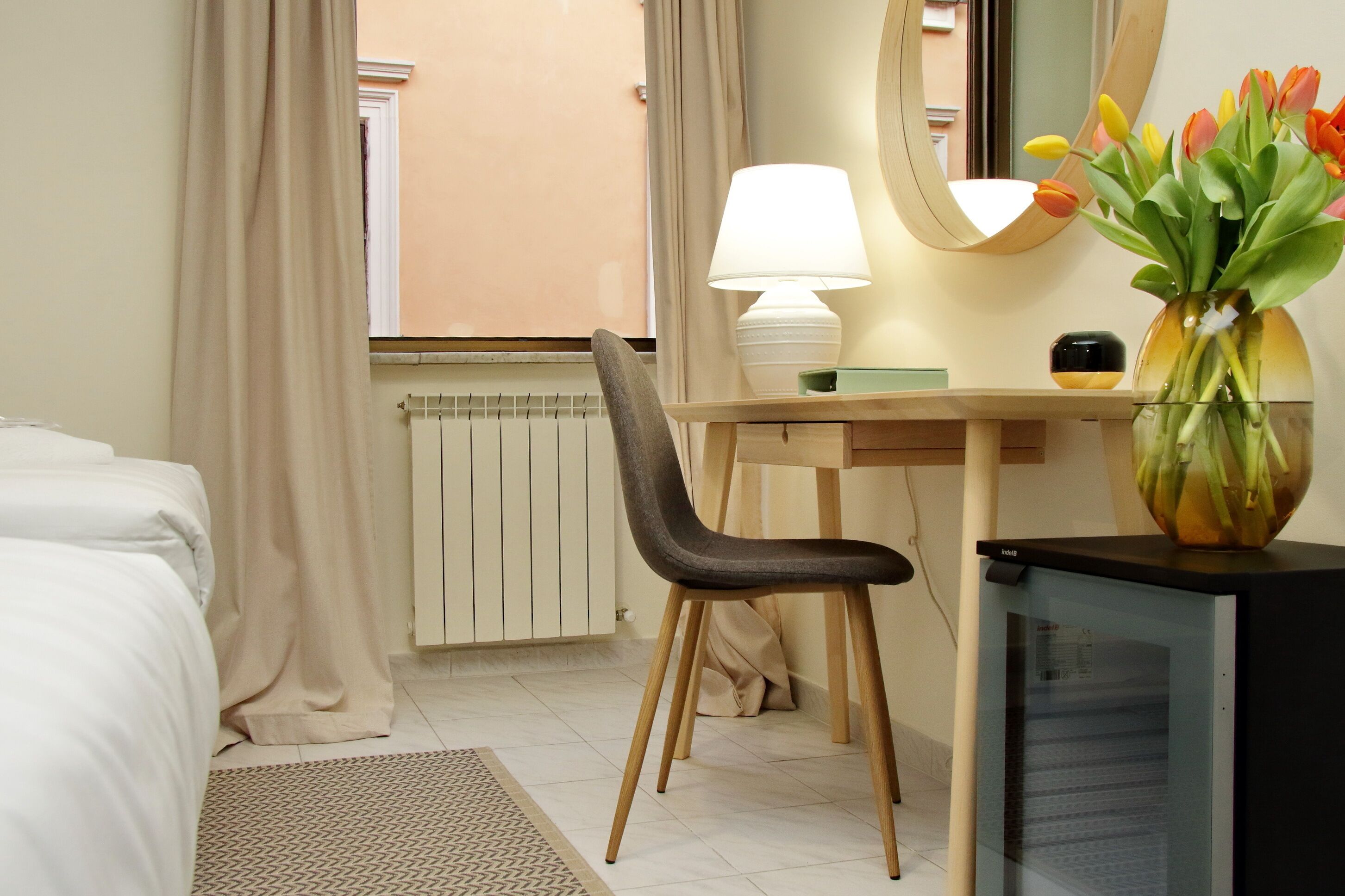 A hotel room with a bed, a desk with a chair, a lamp, a mirror, a vase with flowers, and a radiator.