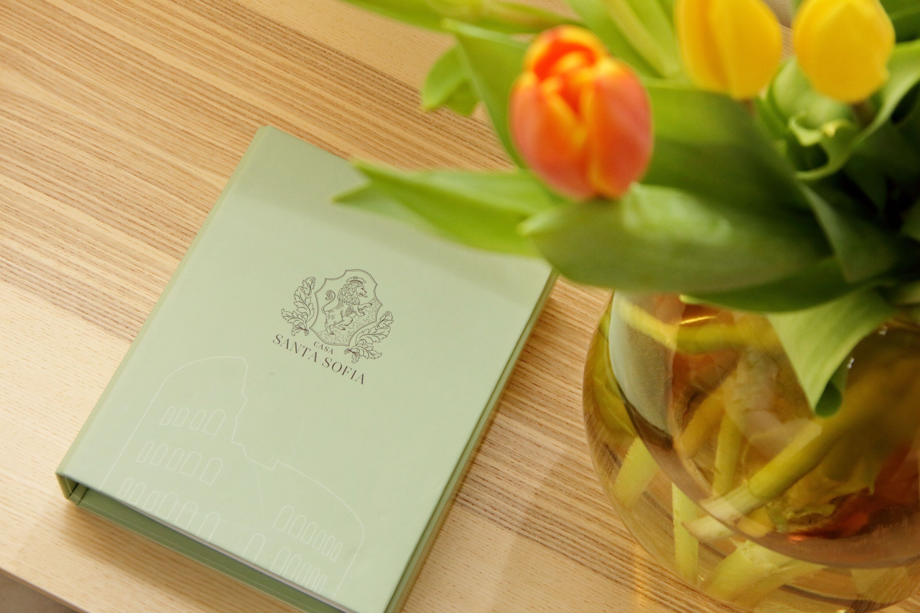 A green book with embossed text on a wooden surface, next to a vase with tulips.
