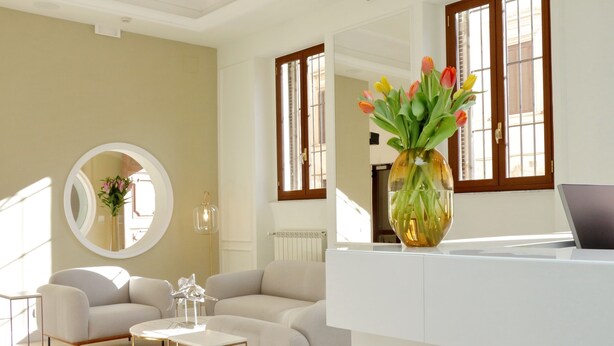 A modern living room with a sofa, coffee table, and a round mirror. There is a vase with flowers on a table, and a laptop on a desk. - Casa Santa Sofia Hotel (Rome)