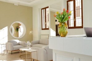 A modern living room with a sofa, coffee table, and a round mirror. There is a vase with flowers on a table, and a laptop on a desk. - Casa Santa Sofia Hotel (Rome)