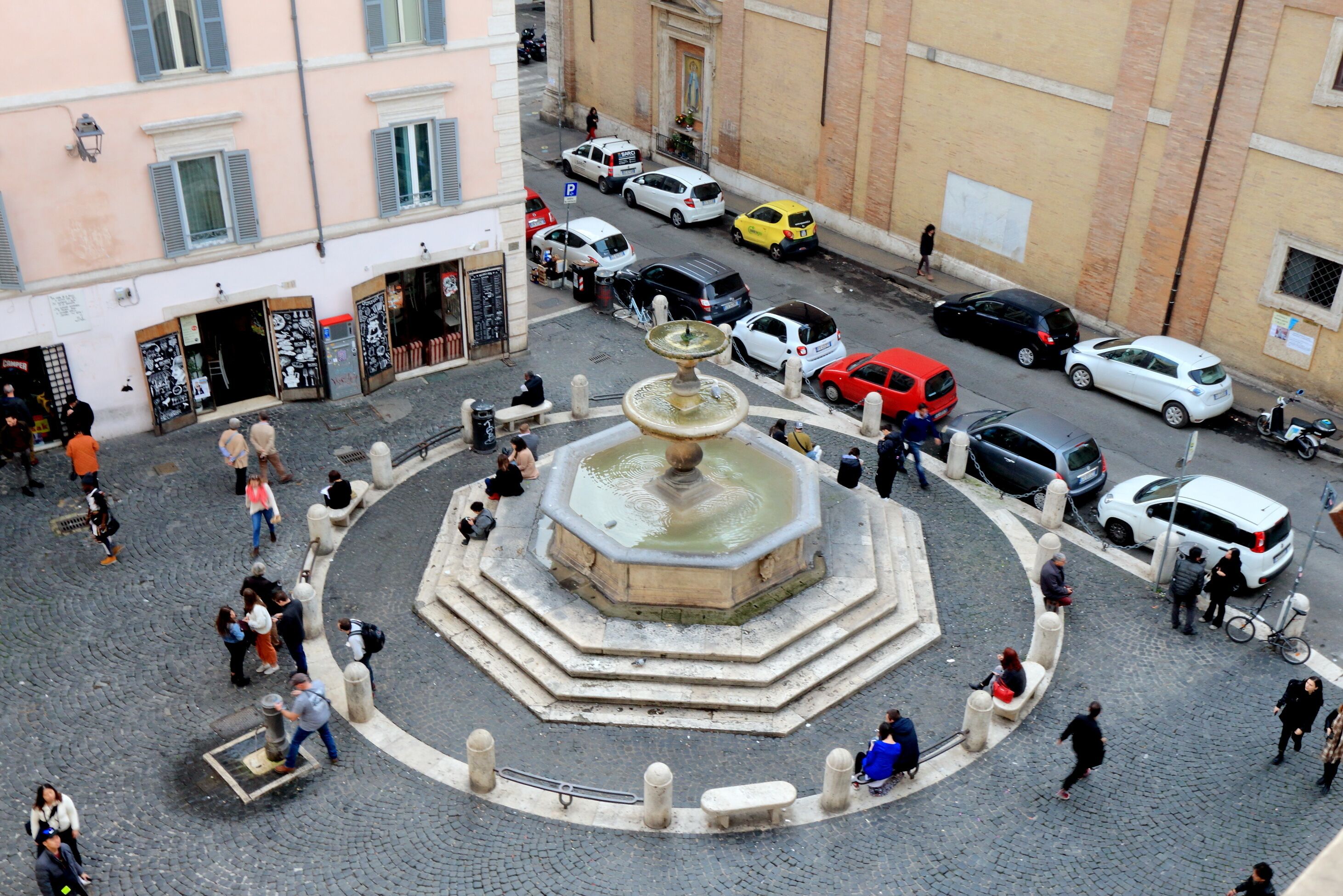 Uma praça pública com uma fonte central, rodeada por calçada de paralelepípedos e diversos edifícios.