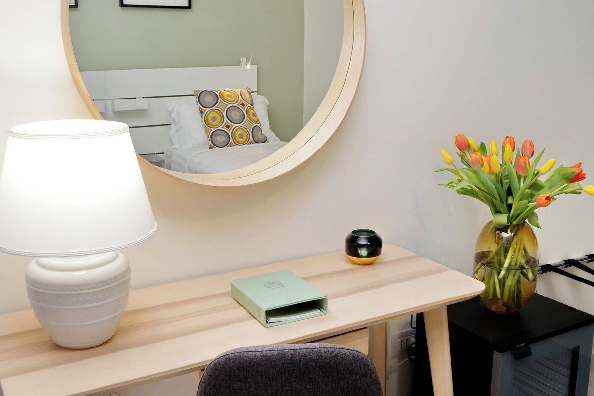 A hotel room with a bed, a bedside table with a lamp, a vase with flowers, and a small green box.