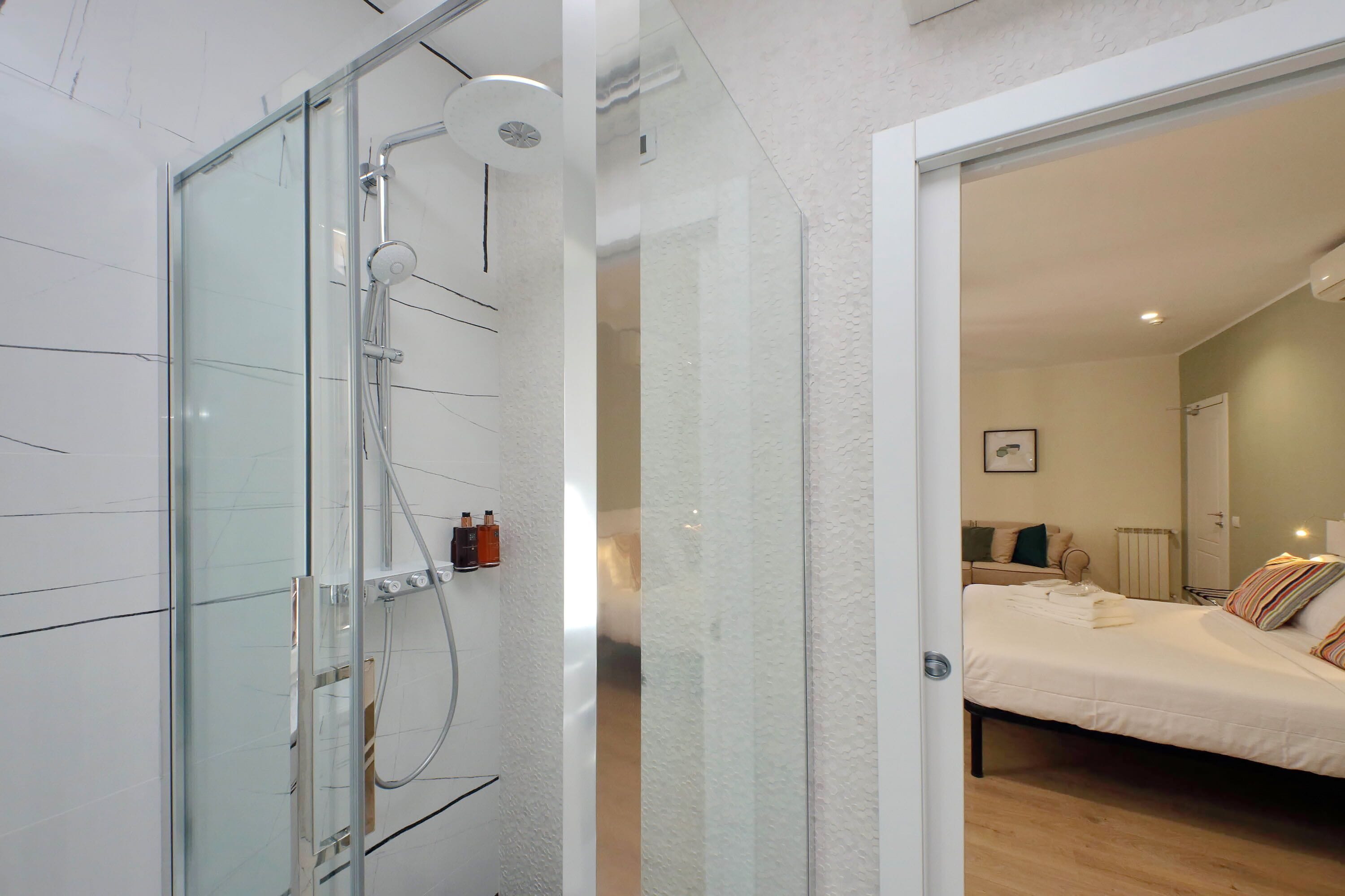 A modern bathroom with a glass shower enclosure, a visible towel rack, and a glass door leading to a bedroom with a bed and a bedside table.