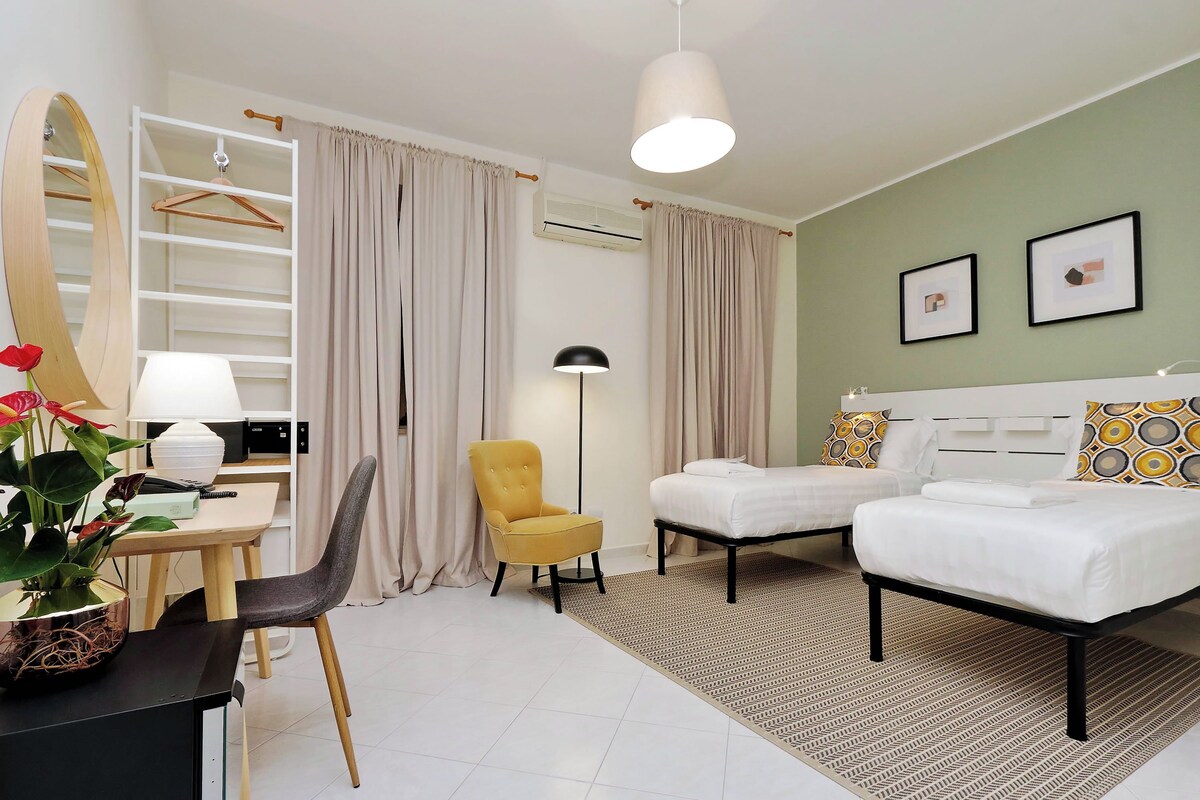 A hotel room with a bed, two bedside tables, a chair, a desk with a lamp, a mirror, a wardrobe, and a large mirror.