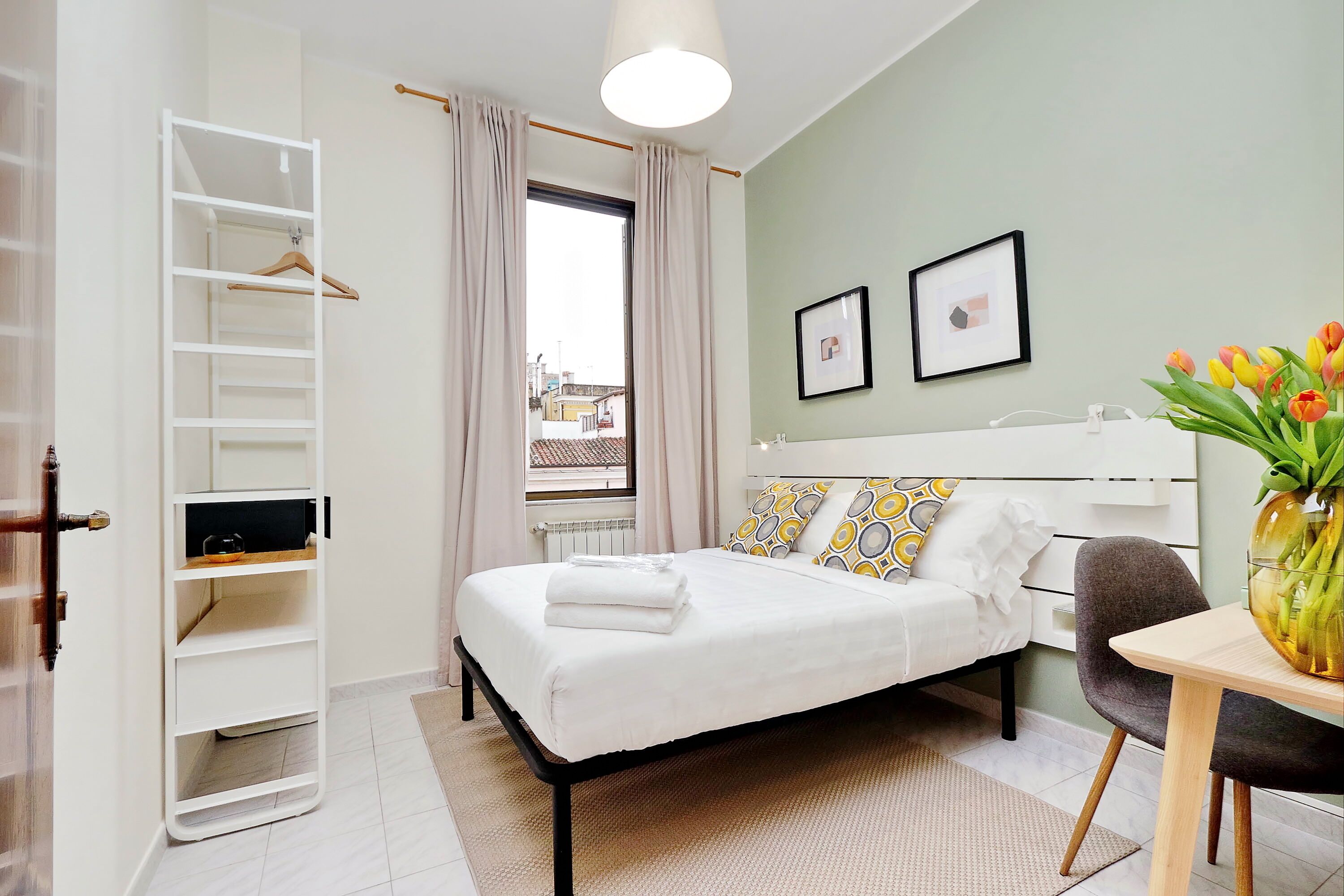 A neatly arranged bedroom with a bed, bedside table, chair, and a wardrobe with a hanging shelf.