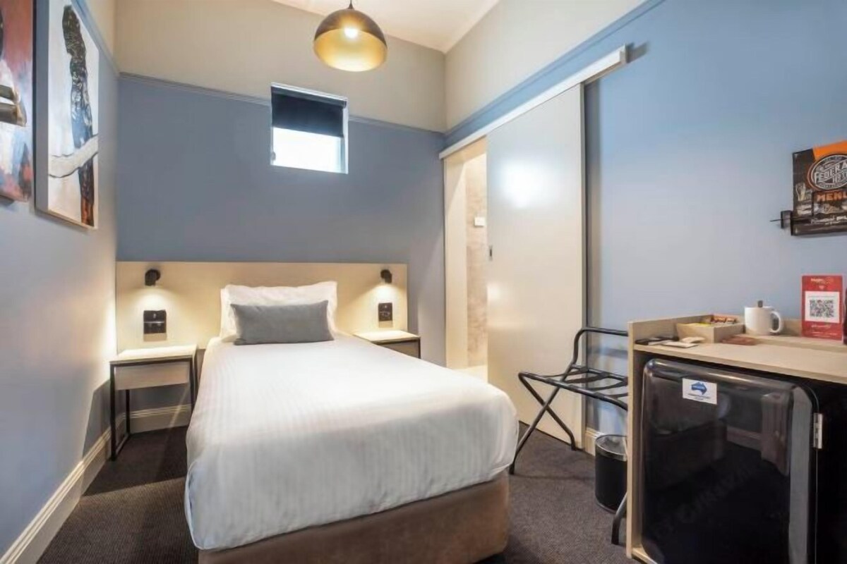 A compact hotel room with a single bed, bedside tables with lamps, a small window, a refrigerator, and a wall-mounted TV.