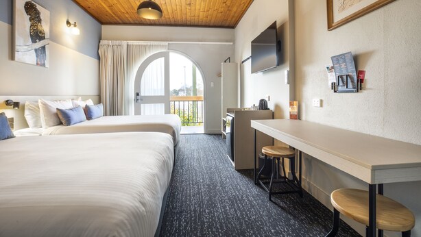 A hotel room with two beds, a desk with stools, a TV, and a large window with a view.