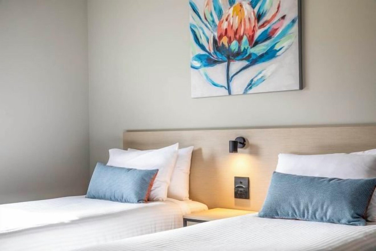 A hotel room with two beds, a nightstand, a wall-mounted lamp, and a floral painting.