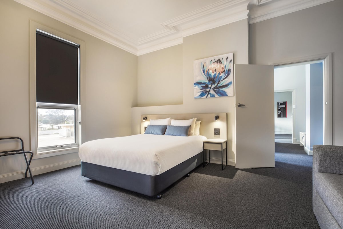 Un cuarto de hotel con una cama grande, una mesita de noche, un cuadro en la pared y vistas por la ventana.