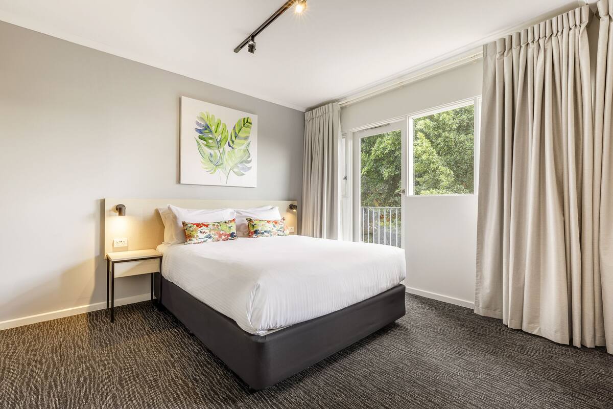 A hotel room with a large bed, a bedside table with a lamp, a window with curtains, and a wall-mounted artwork.