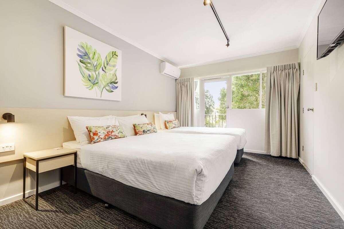 A hotel room with two beds, a nightstand, a wall-mounted air conditioner, and a window with curtains.