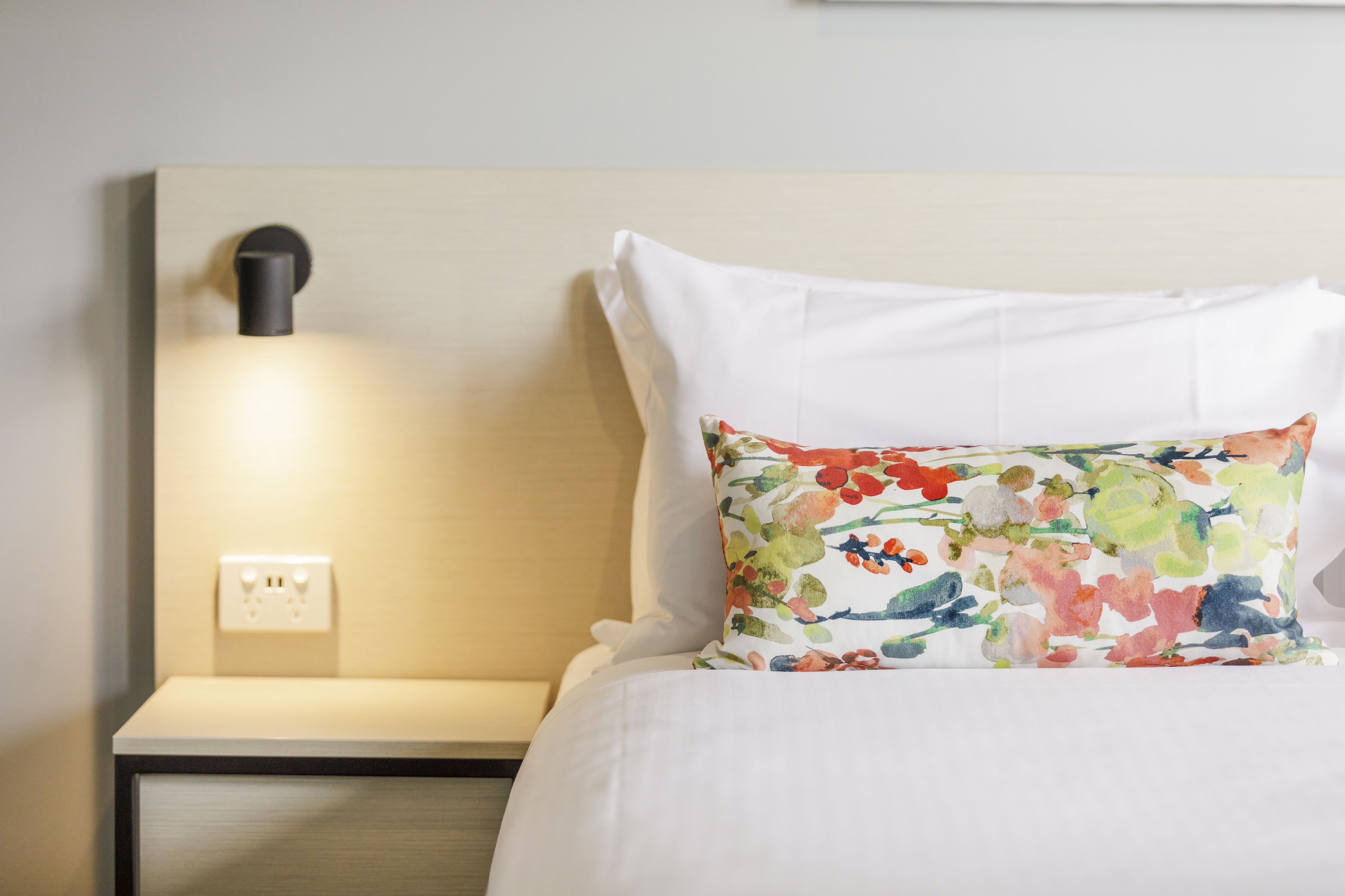 A neatly made bed with white linens and a colorful floral pillow. There is a bedside table with a lamp and a power outlet.