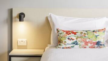 A neatly made bed with white linens and a colorful floral pillow. There is a bedside table with a lamp and a power outlet.