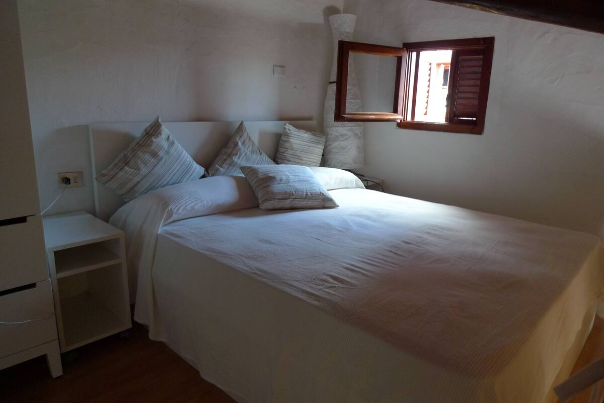 A neatly made bed with white linens and multiple pillows, a small bedside table, and a wall-mounted mirror.