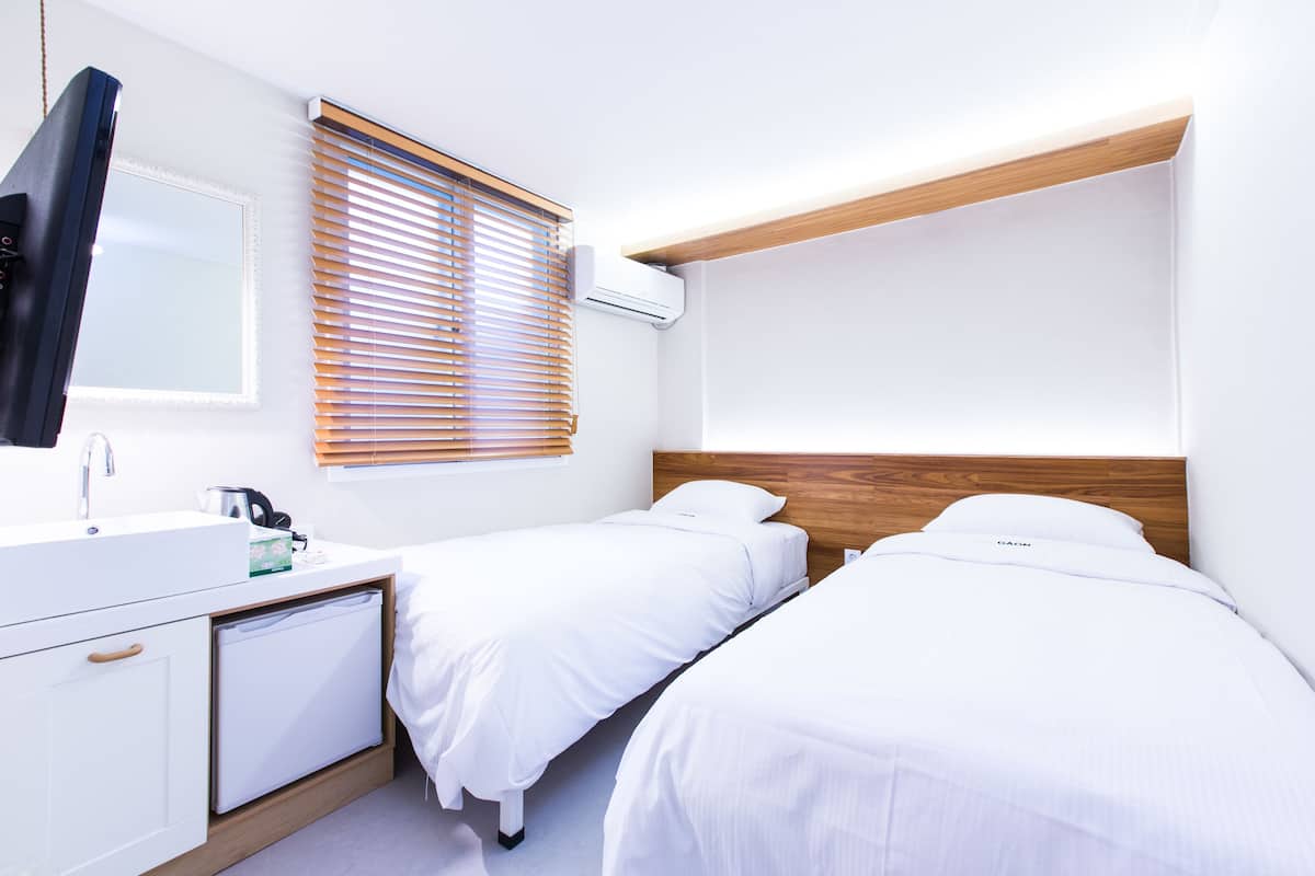 A hotel room with two single beds, a built-in TV, a sink, and a window with blinds.