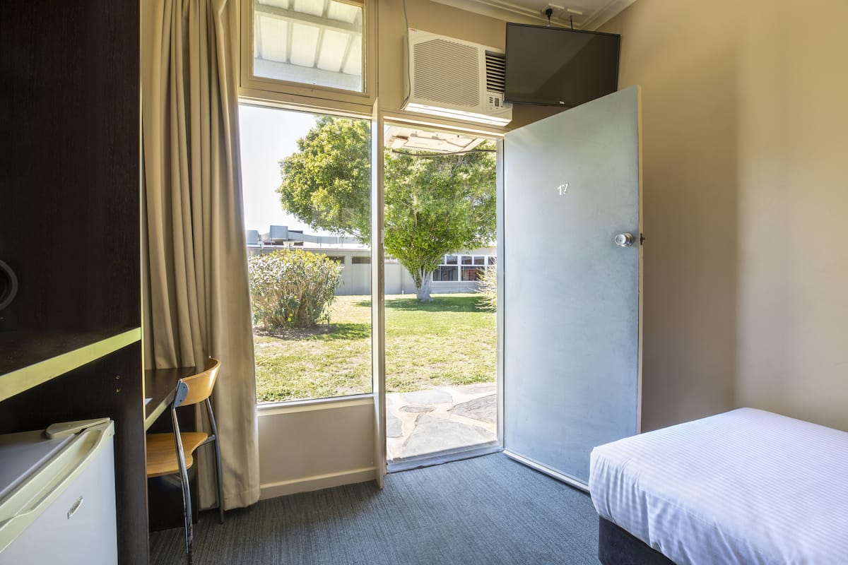 A hotel room with a bed, a desk with a chair, a window with curtains, and an air conditioning unit.