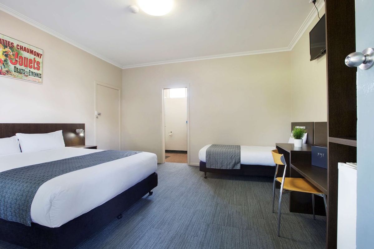 A hotel room with two beds, a desk with a chair, a TV, and a door leading to another room.