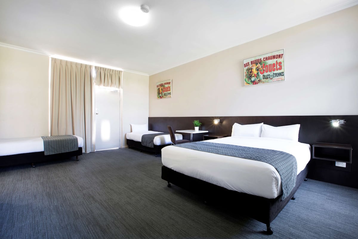 A hotel room with a large bed, two smaller beds, a desk, and a chair. There are posters on the wall and a plant on the desk.