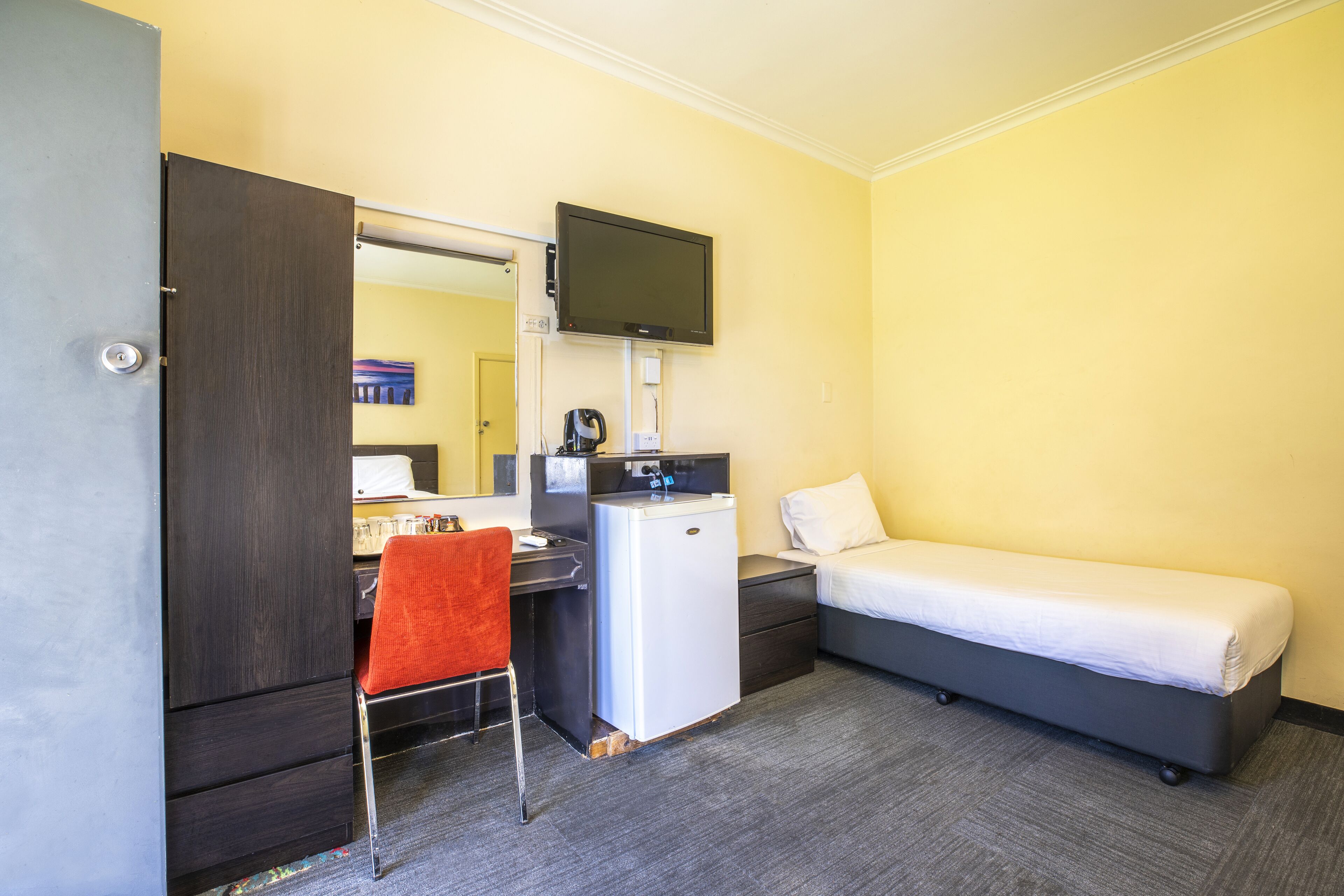 A compact hotel room with a bed, a desk with a chair, a small refrigerator, a television mounted above the desk, and a mirror on the wall.