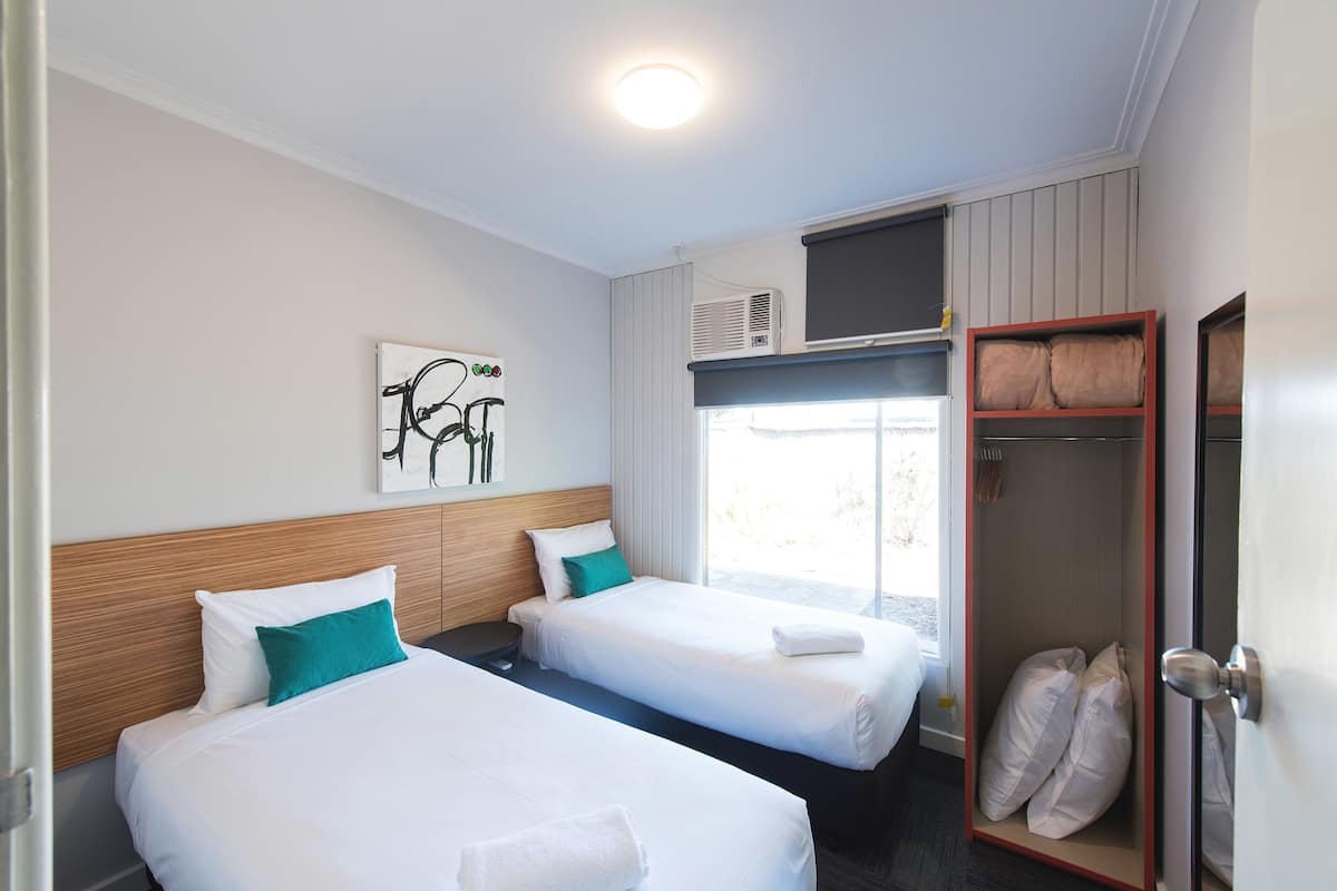 A hotel room with two beds, a window with blinds, a ceiling light, and a wall-mounted air conditioning unit.