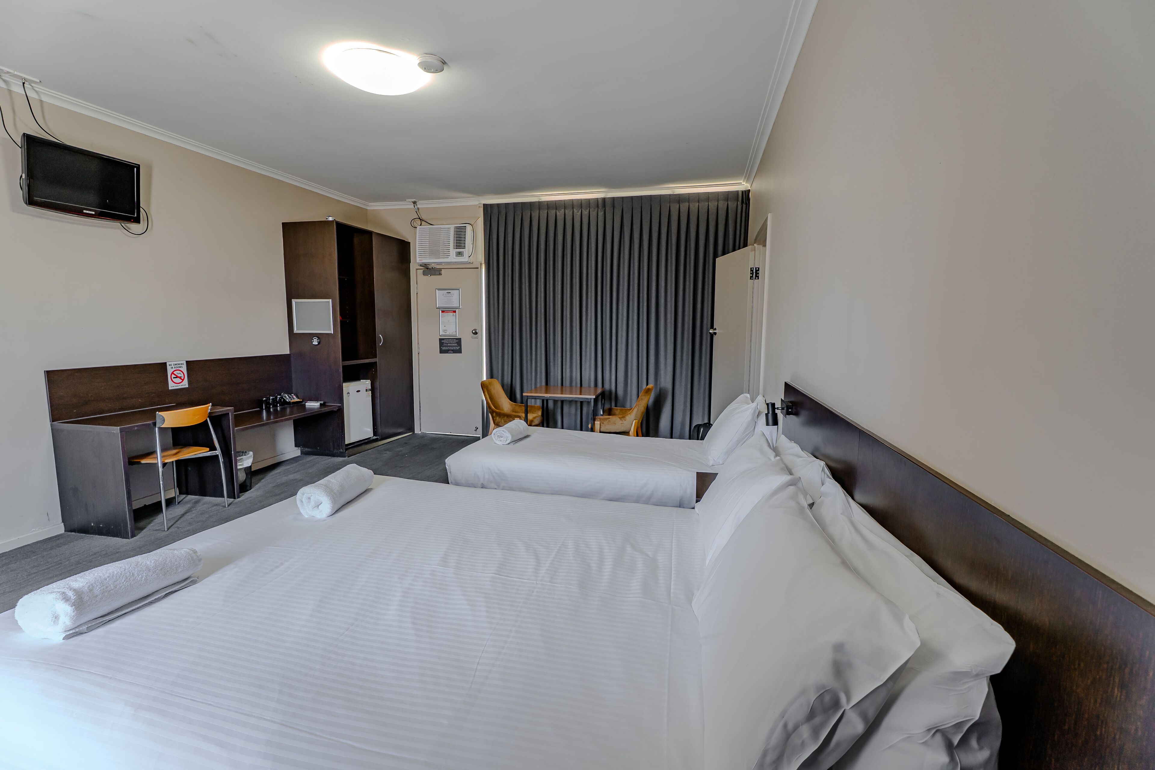 A hotel room with two beds, a desk, a chair, a TV, and an air conditioning unit.