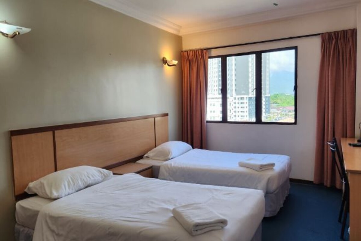 Habitación de hotel con dos camas individuales, cabecera de madera, ventana grande con cortinas y vistas a edificios.