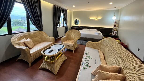 A hotel room with a bed, two wicker chairs, a small table with a remote control, and a view of the outdoors through large windows.