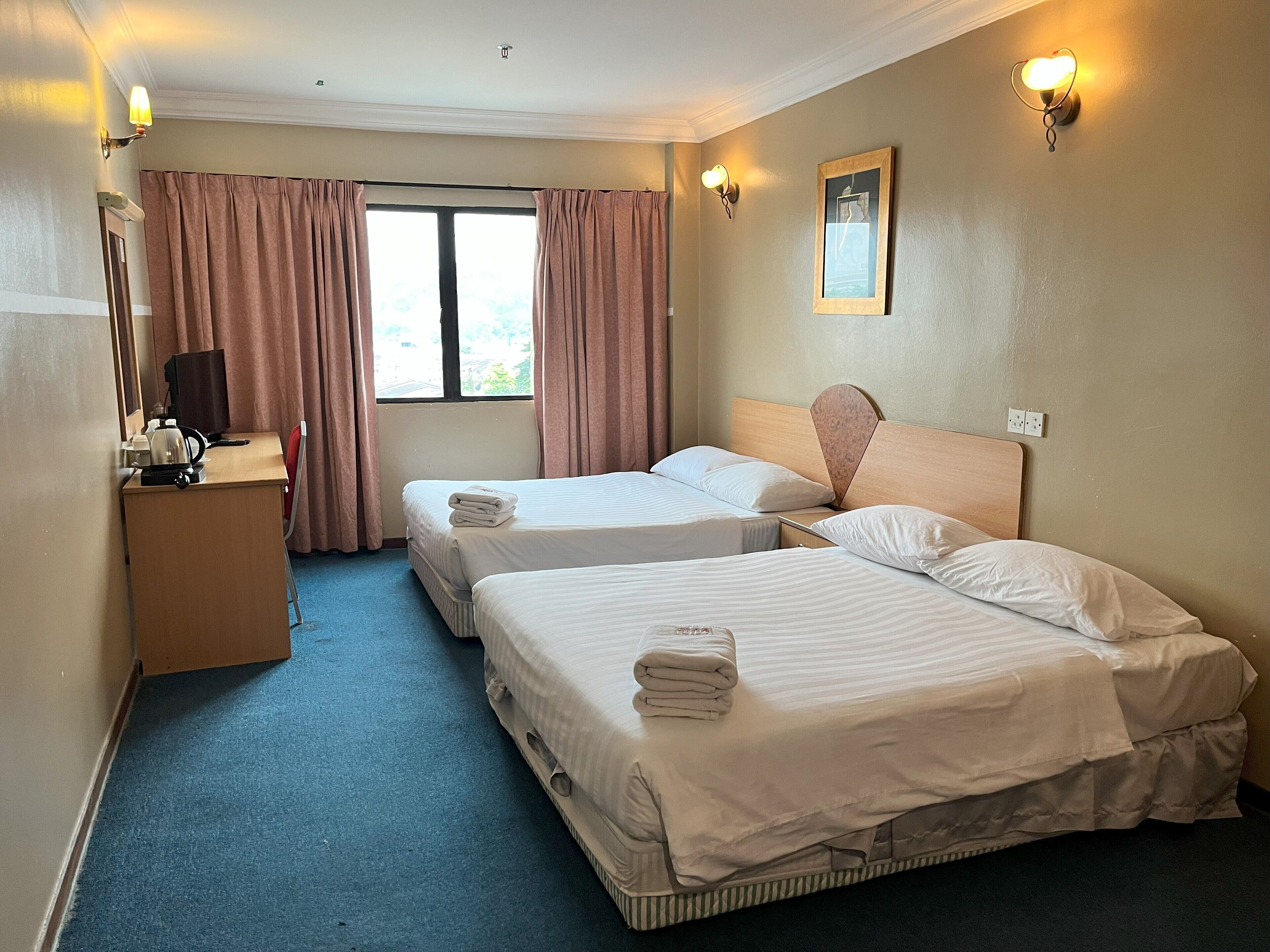 A hotel room with two beds, a desk with a kettle, a chair, and a window with curtains.