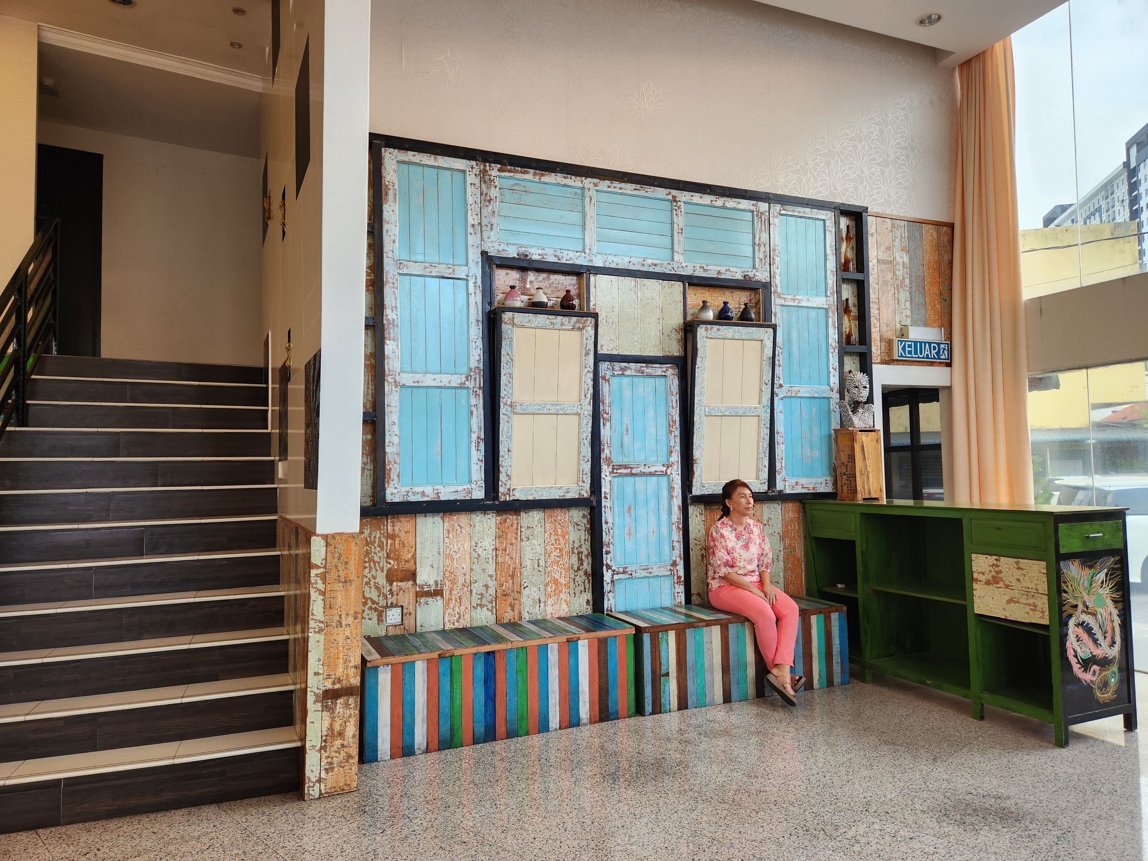 Une femme est assise sur un banc coloré dans un hall d’entrée, près d’un escalier et d’un comptoir vert.