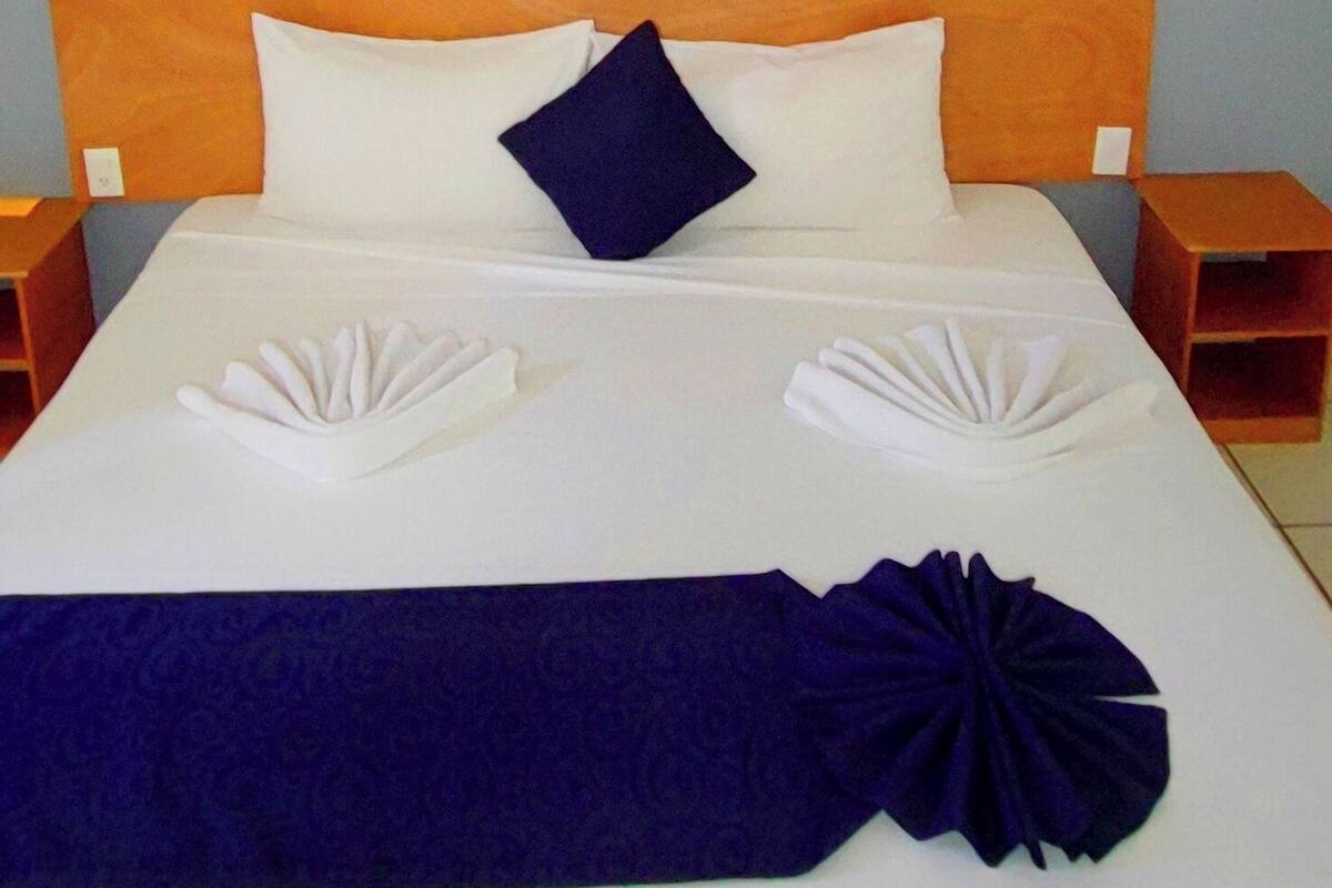 Una cama bien hecha con una colcha blanca y azul, dos almohadas blancas y una almohada azul oscuro, junto con un arreglo decorativo de toallas azules y blancas.