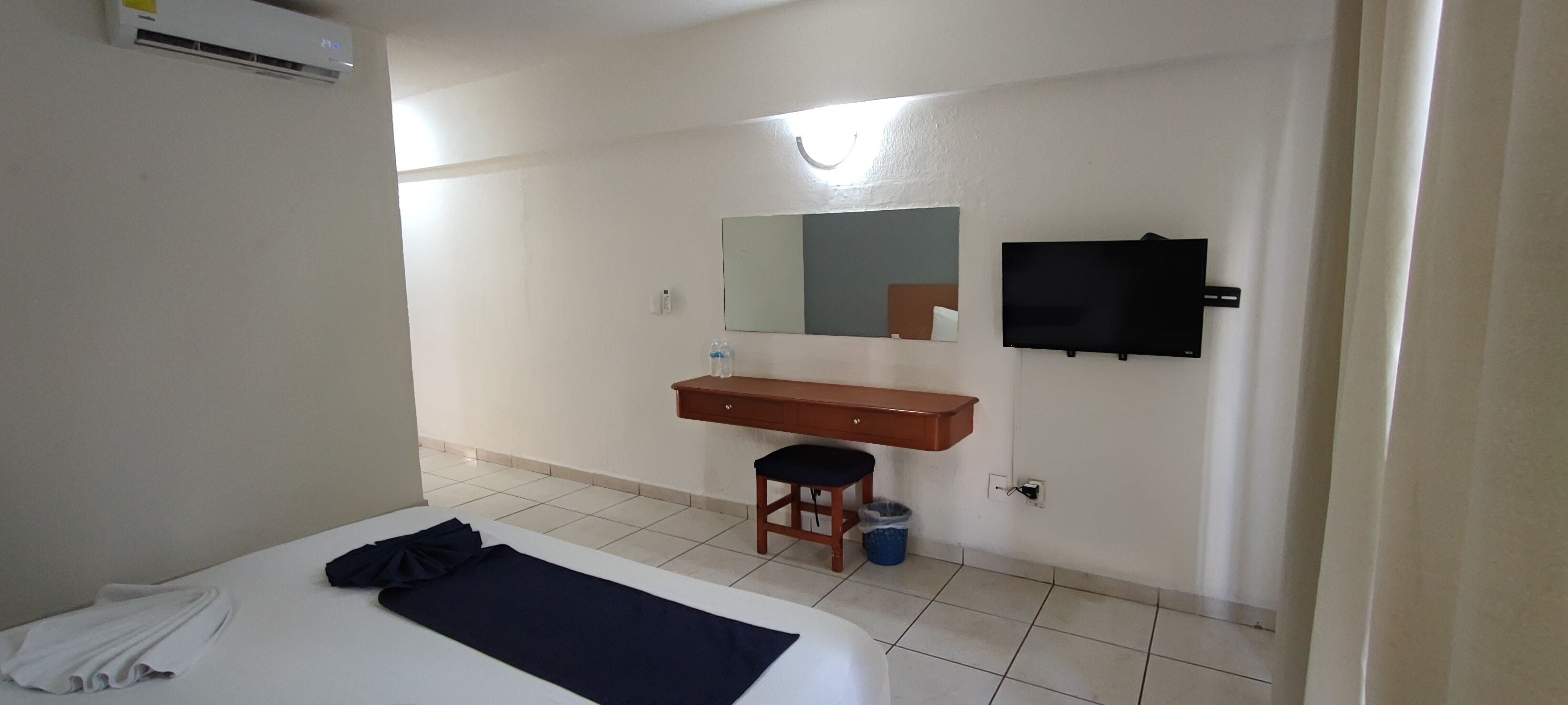 Quarto de hotel com cama, cômoda de madeira, espelho, televisão e um banquinho.