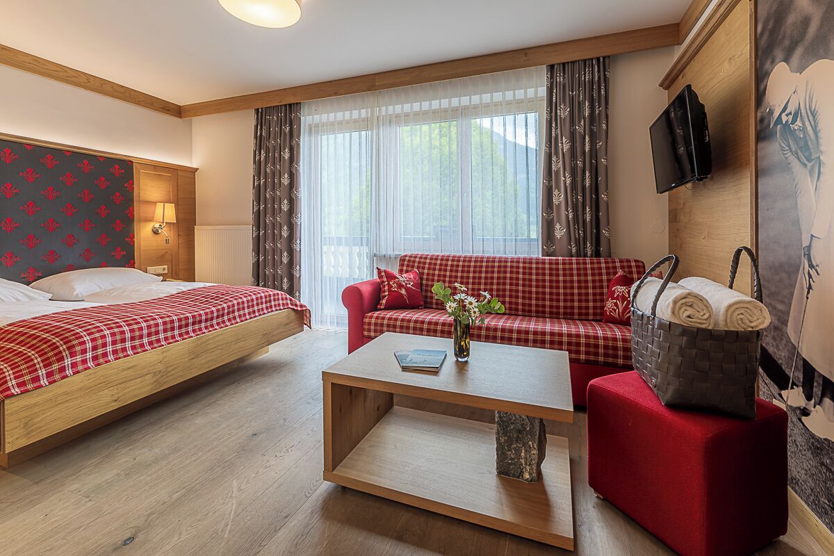 Habitación de hotel con cama, sofá, mesa de centro, televisor y ventana con cortinas.