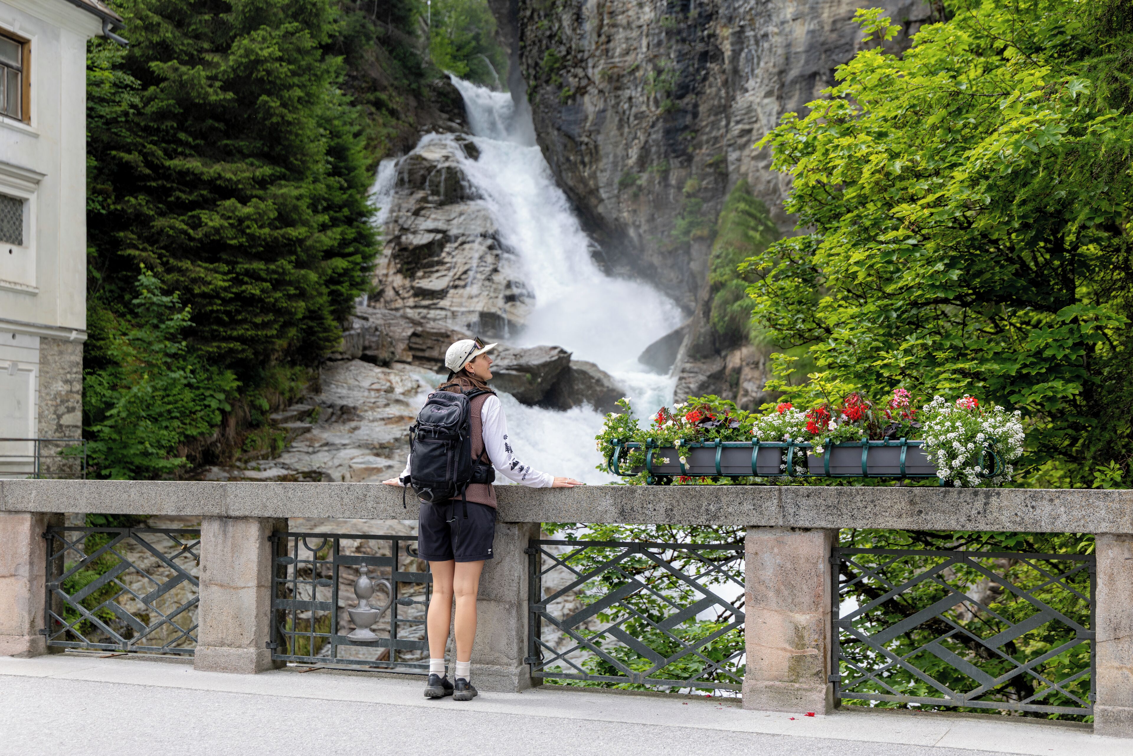 A person standing by a railing overlooking a waterfall, with greenery and flowers in the foreground.