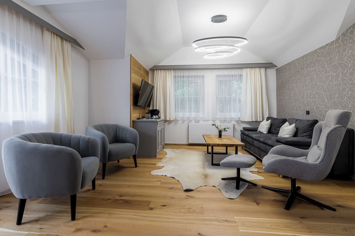 A modern living room with a wooden floor, grey upholstered chairs, a black sofa, a small table, and a mounted TV.