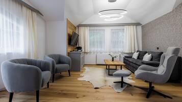 A modern living room with a wooden floor, grey upholstered chairs, a black sofa, a small table, and a mounted TV.