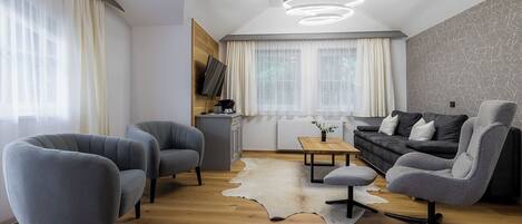 A modern living room with a wooden floor, grey upholstered chairs, a black sofa, a small table, and a mounted TV.