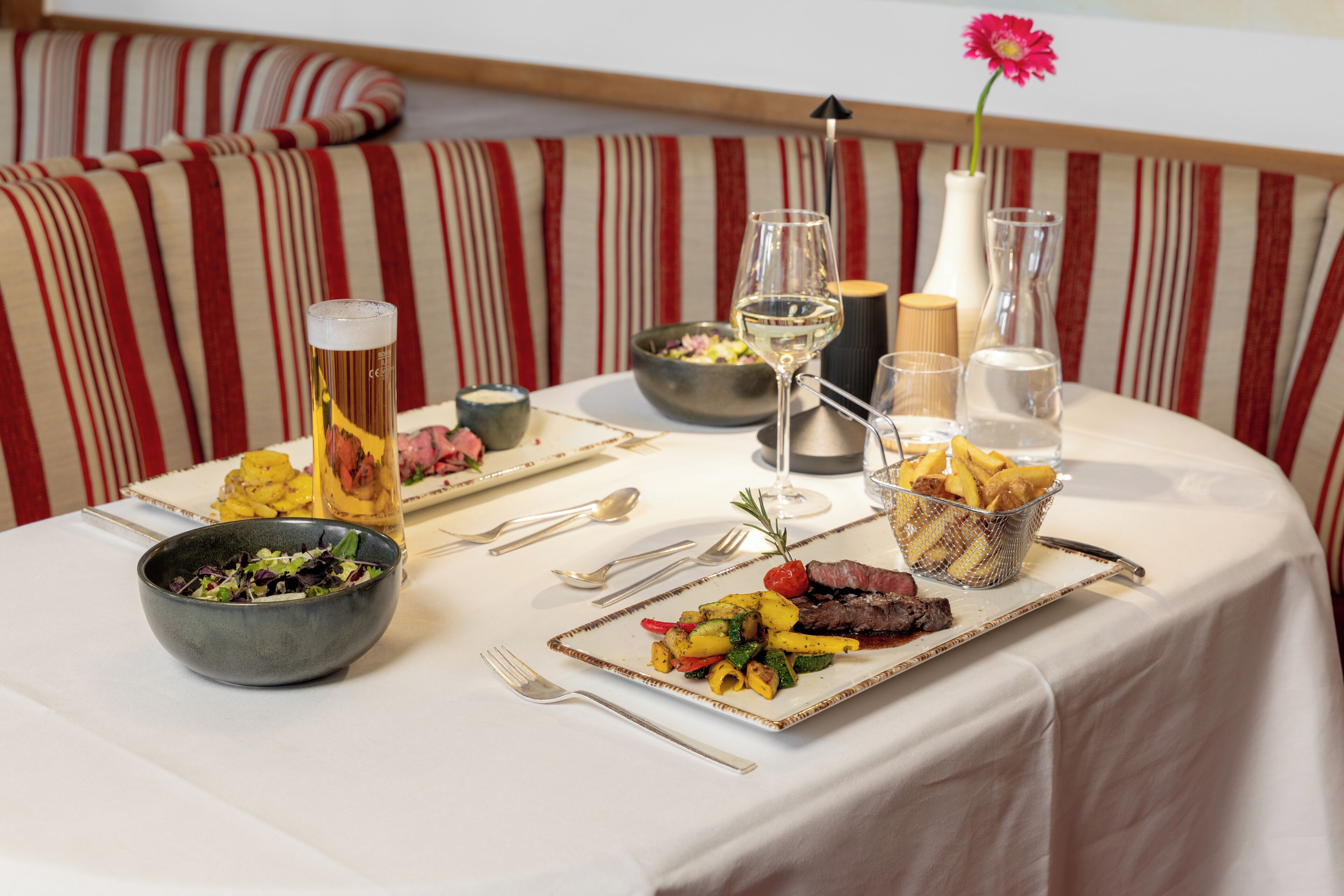 A dining table set with a plate of food, a glass of beer, a bowl of salad, and a vase with a flower.