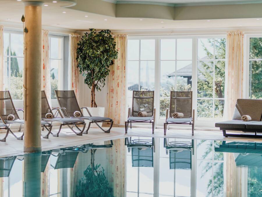 Una piscina interior con sillones de descanso y un sofá, ventanas amplias con cortinas y una planta en maceta.