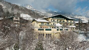 Ein mehrstöckiges Hotel mit verschneitem Dach, umgeben von schneebedeckten Bäumen und Bergen im Hintergrund.