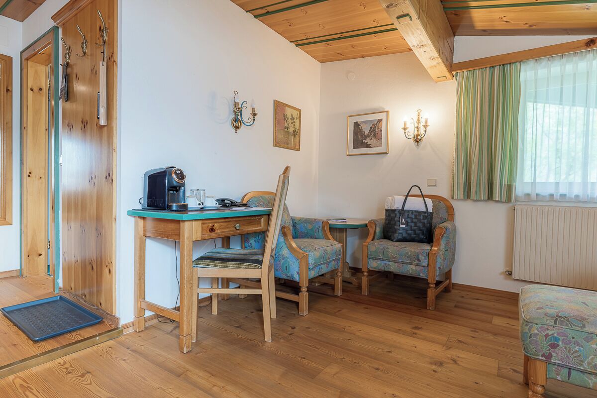 A cozy room with wooden floors, a dining table with chairs, a sofa, a small table with a lamp, and a window with curtains.