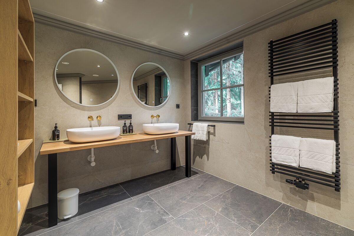 Een moderne badkamer met twee wastafels, een grote spiegel, een handdoekrek en een raam met uitzicht op bomen.