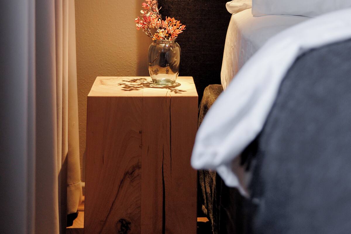Una cama bien tendida con sábanas blancas, una mesita de noche de madera con un florero y una lámpara de pared.