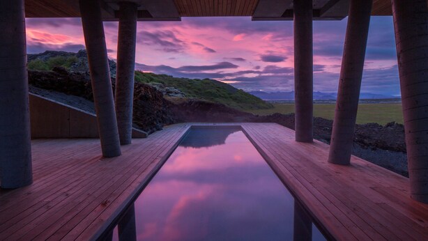 A wooden deck with a pool reflecting a sunset sky, surrounded by columns and a view of a hilly landscape. - ION Adventure Hotel, Nesjavellir, a Member of Design Hotels (Selfoss)