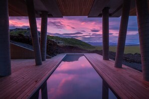 Una terraza de madera con piscina que refleja un cielo al atardecer, rodeada de columnas y vistas a un paisaje montañoso.