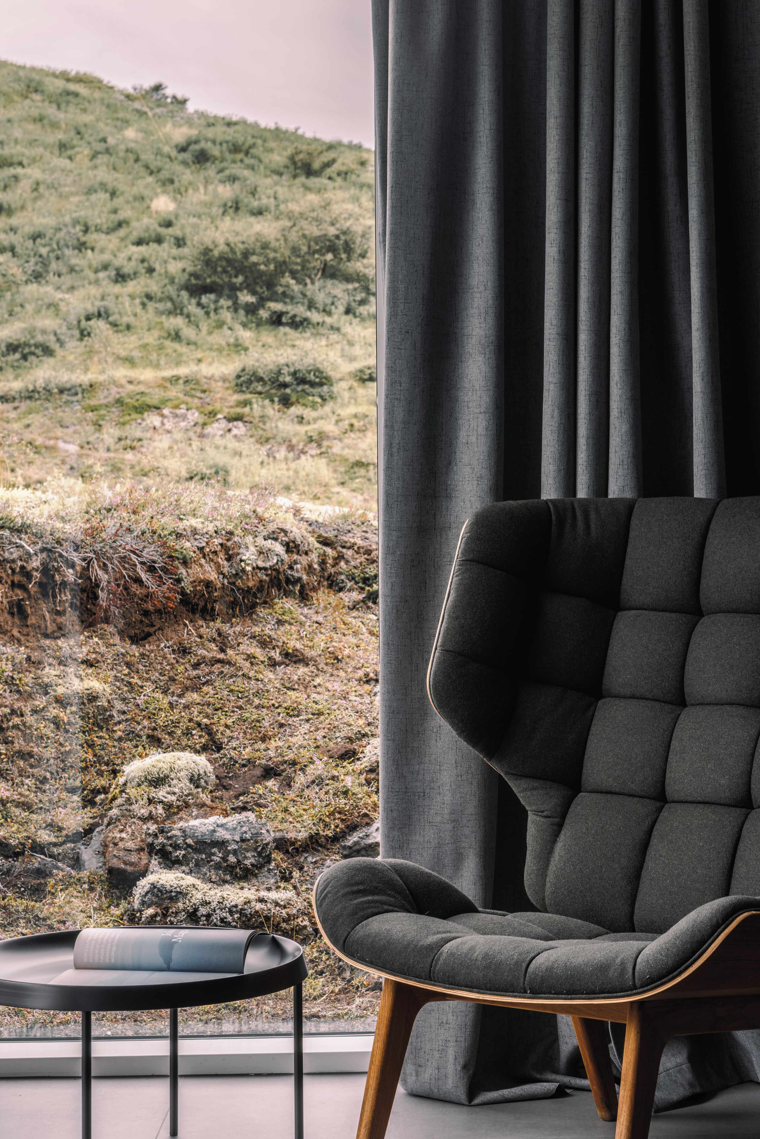 A modern room with a view of a rocky hillside, a dark grey upholstered chair, and a small black table.