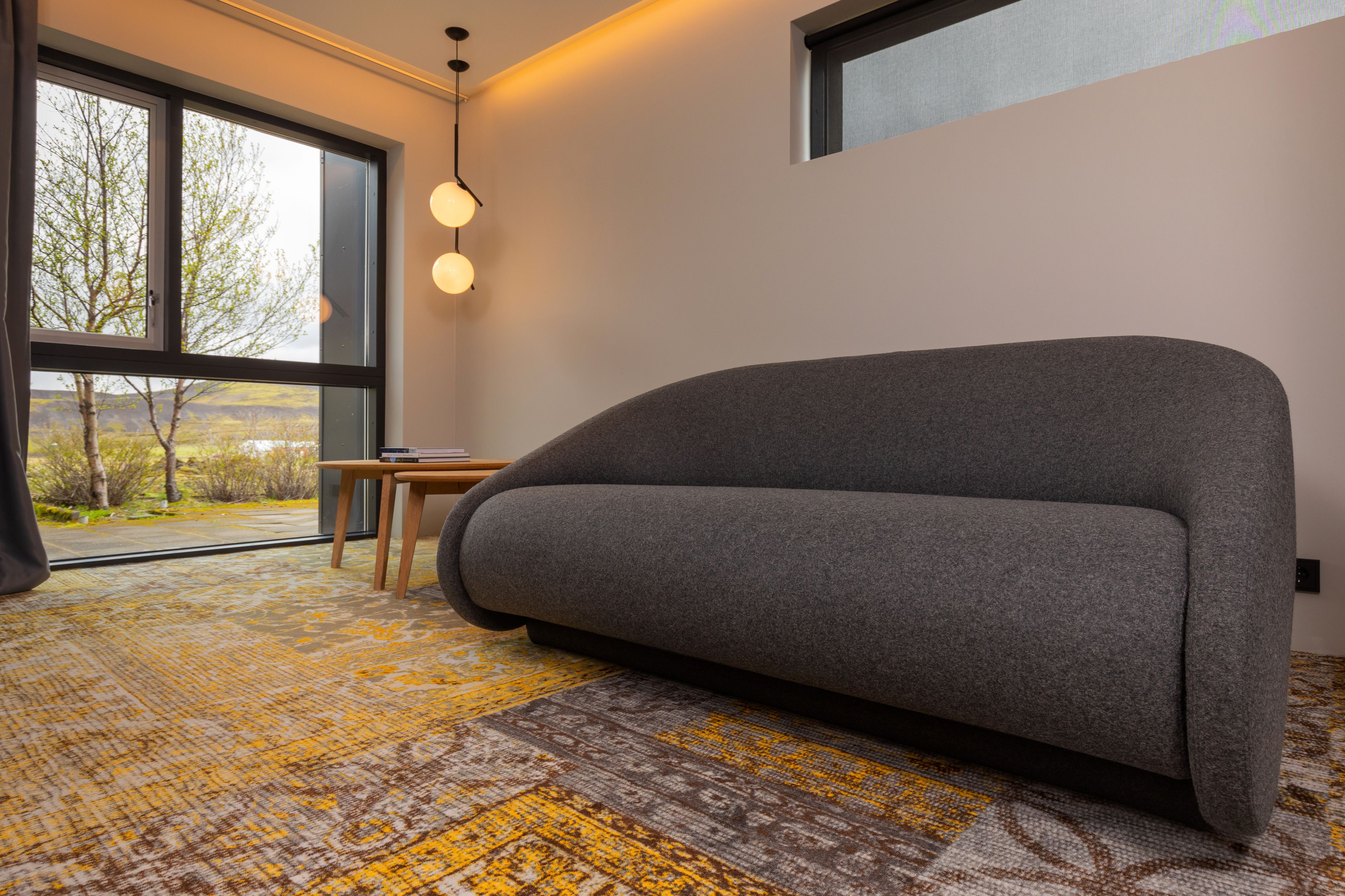 A modern living room with a grey sofa, a wooden side table, and a large window offering a view of trees and hills.