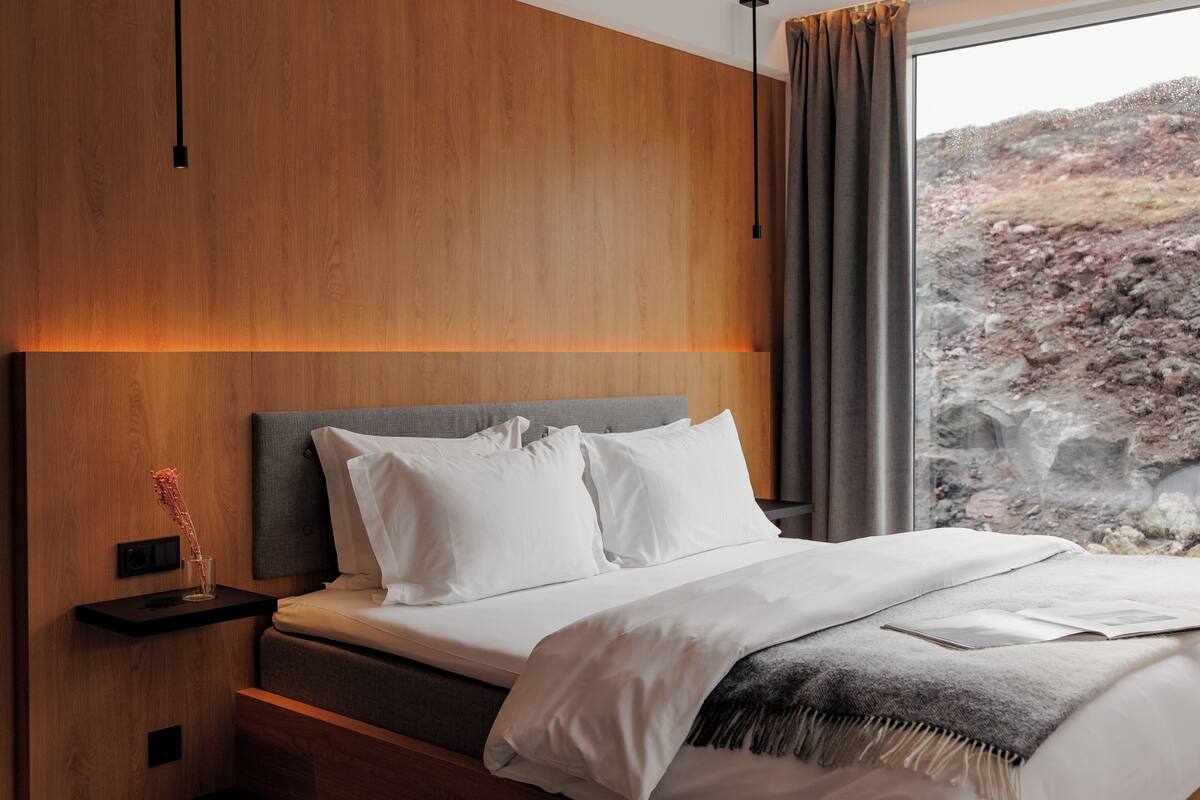 Un dormitorio moderno con cabecera de madera, una mesita de noche con lámpara y vistas a un paisaje rocoso a través de la ventana.