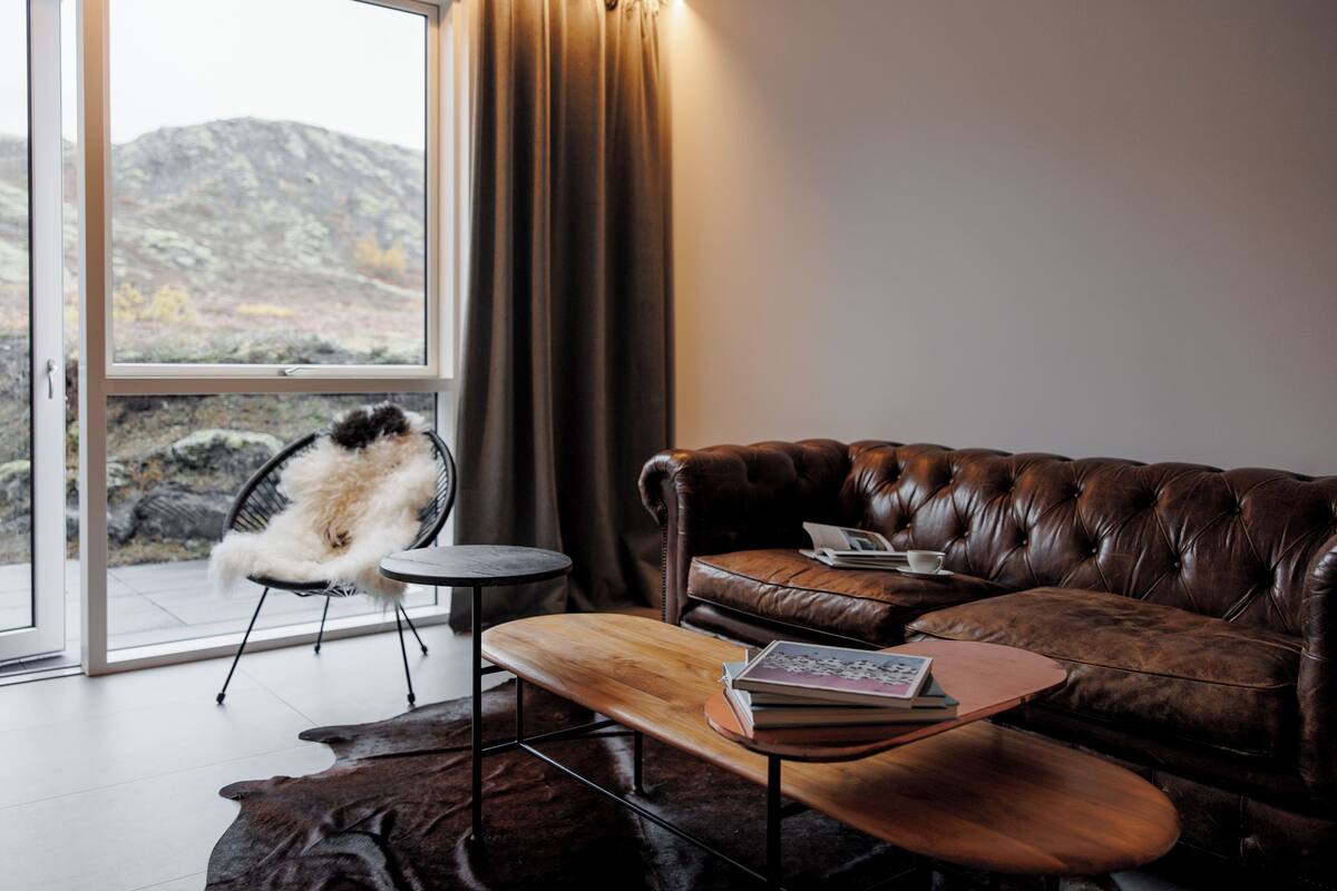 Un salón moderno con un sofá de cuero, una mesa de centro de madera y un sillón blanco mullido junto a un ventanal con vistas a un paisaje rocoso.