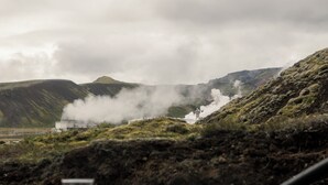 A table with a glass and a bottle, a chair, and a view of a mountainous landscape with a building emitting steam. - ION Adventure Hotel, Nesjavellir, a Member of Design Hotels (Selfoss)