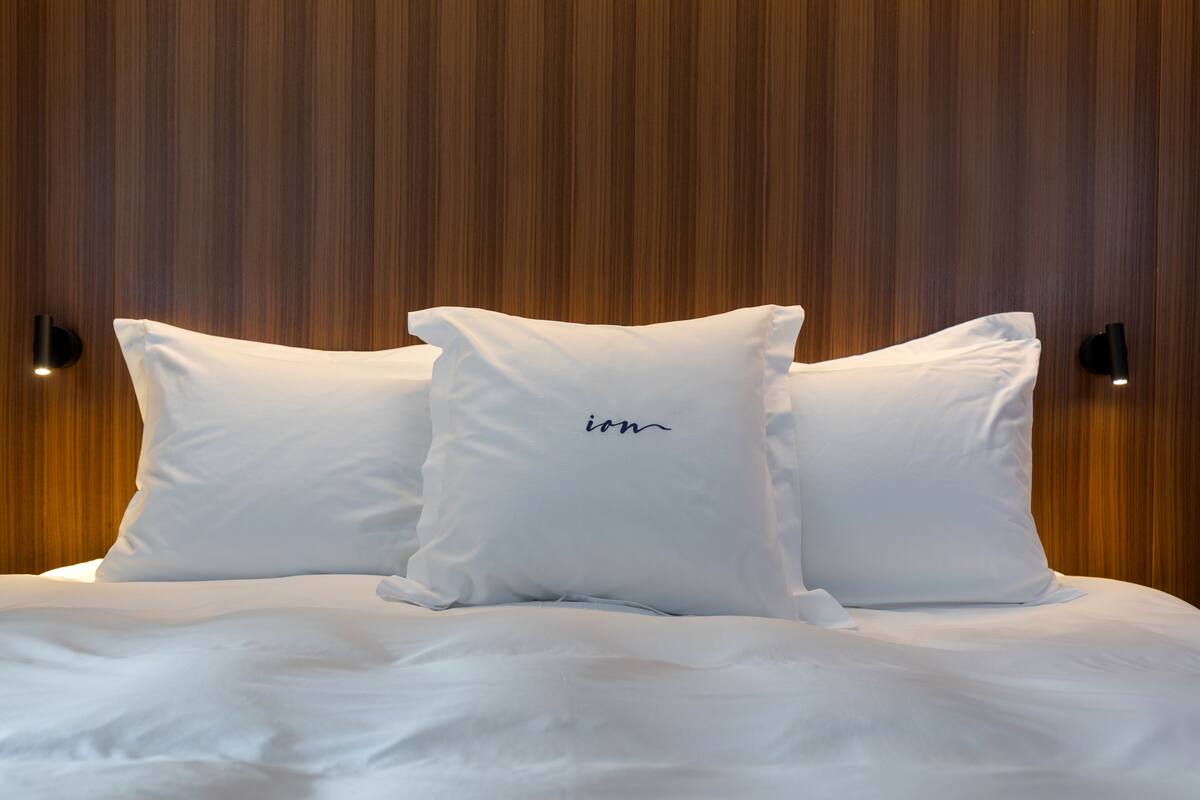 Una cama con ropa de cama y almohadas blancas, cabecera de madera y dos lámparas fijadas a la pared.